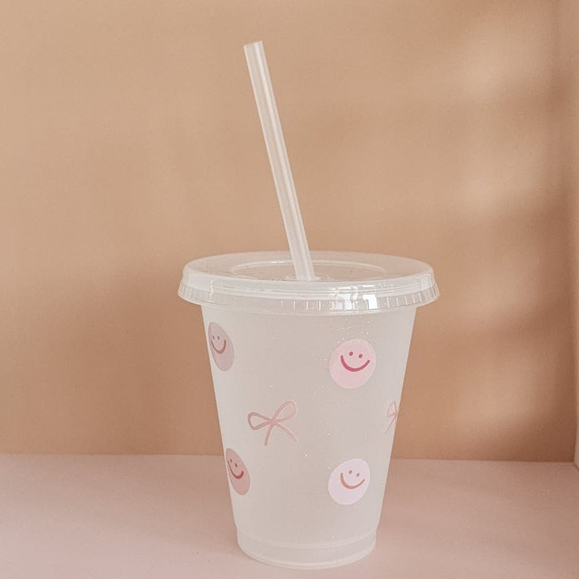 Trinkbecher aus Kunststoff mit Strohhalm, verziert mit rosa Smiley- und Schleifenmotiven, auf hellem Hintergrund