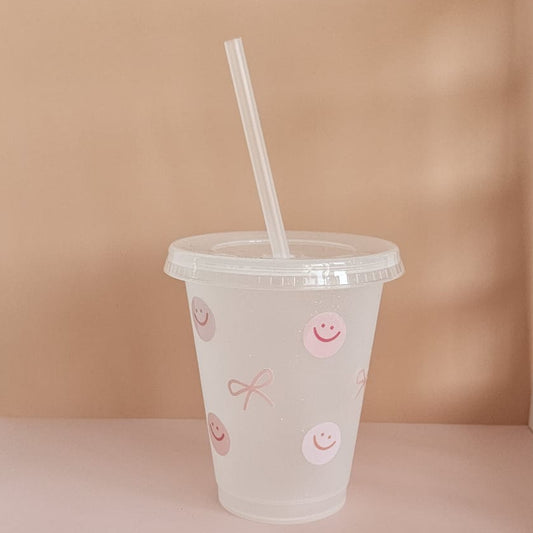 Trinkbecher aus Kunststoff mit Strohhalm, verziert mit rosa Smiley- und Schleifenmotiven, auf hellem Hintergrund